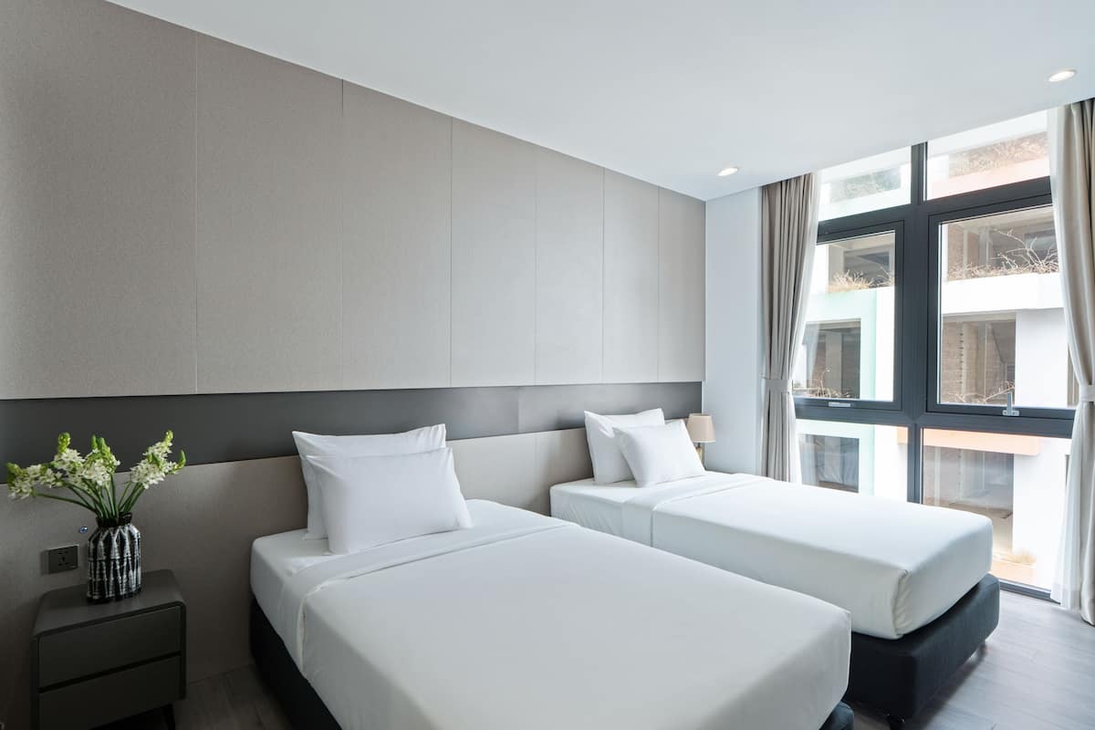 A hotel room with two beds, a nightstand, a vase with flowers, and a large window with curtains.