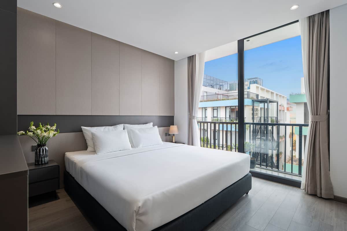 A modern hotel room with a large bed, a nightstand with a vase of flowers, a window with a view of buildings, and a balcony with a railing.
