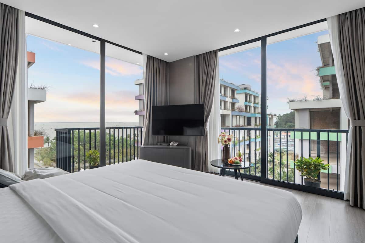 A modern hotel room with a large bed, a flat-screen TV, and a balcony with a view of the cityscape.