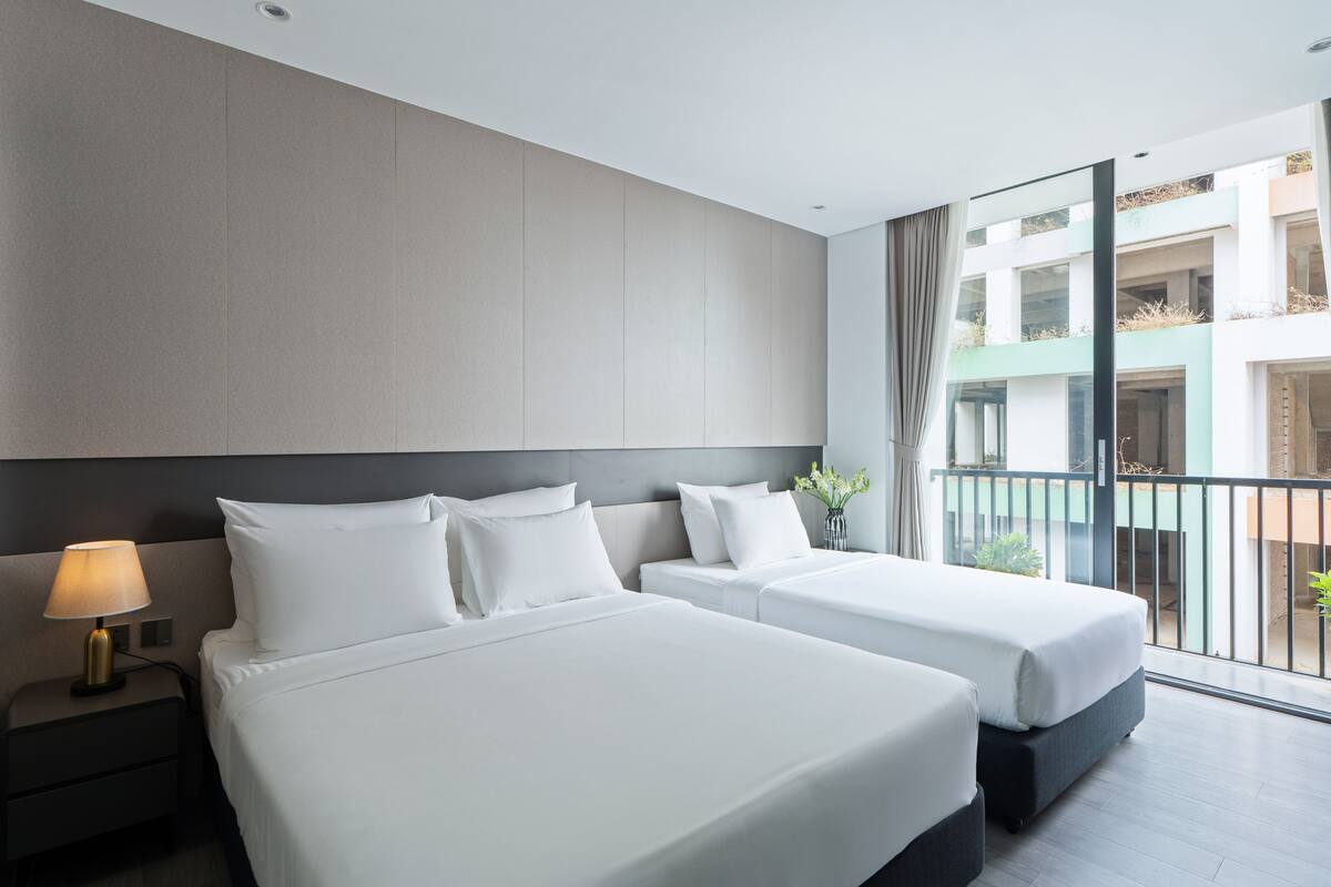A hotel room with two beds, a nightstand with a lamp, a large window with a view of the building's exterior, and a balcony with plants.