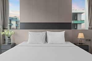Quarto de hotel com uma cama grande, duas almofadas, mesa de cabeceira com abajur, um vaso com flores e vista para edifícios pela janela.