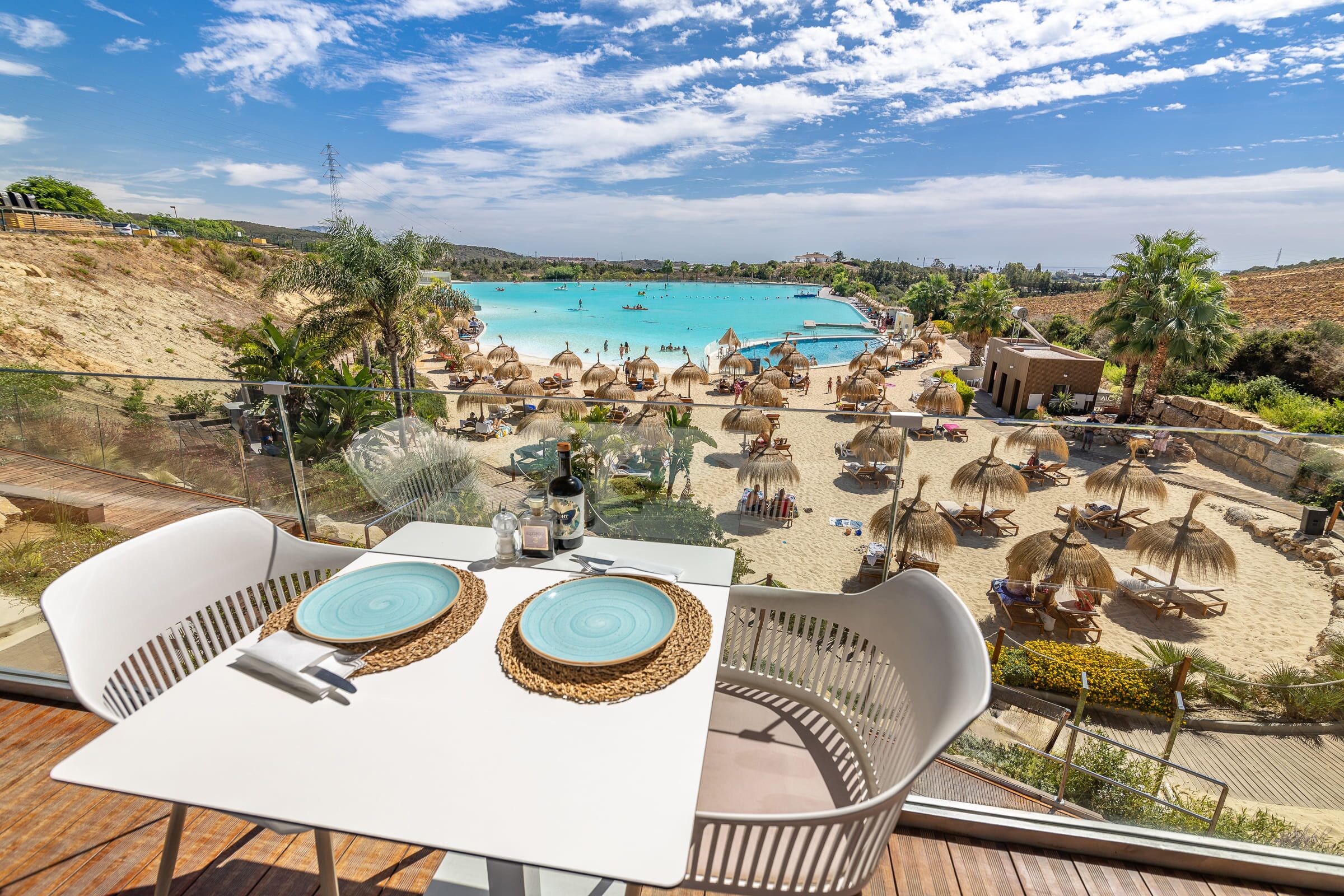 Eine Terrasse mit Tisch für zwei Personen, mit Blick auf einen Strand mit Sonnenschirmen und Liegestühlen.