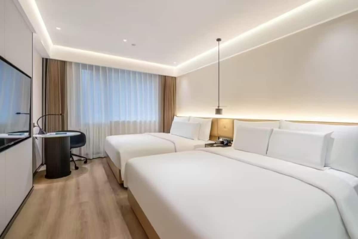 A hotel room with two beds, a desk, a chair, a lamp, and a large window with curtains.