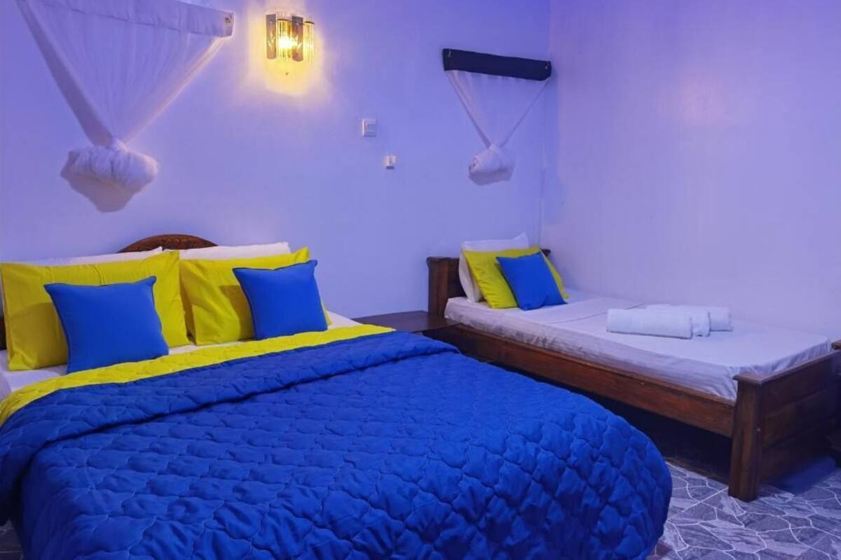 A room with two beds, each with blue and yellow bedding. There are wall-mounted lights and a mosquito net above one bed.