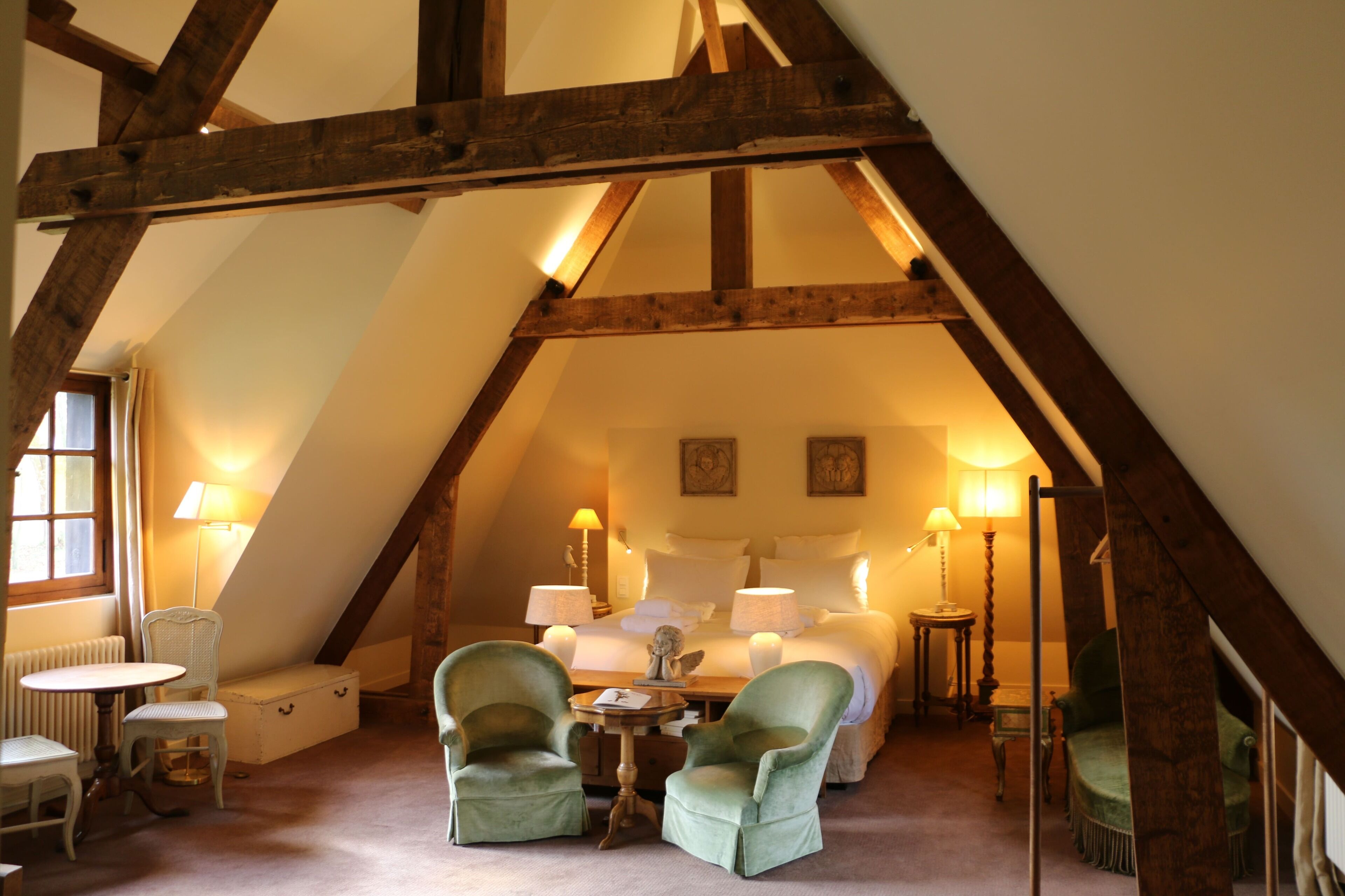 Une chambre mansardée chaleureuse avec un plafond en pente, un lit avec du linge de maison blanc, deux fauteuils verts, une petite table et une lampe.