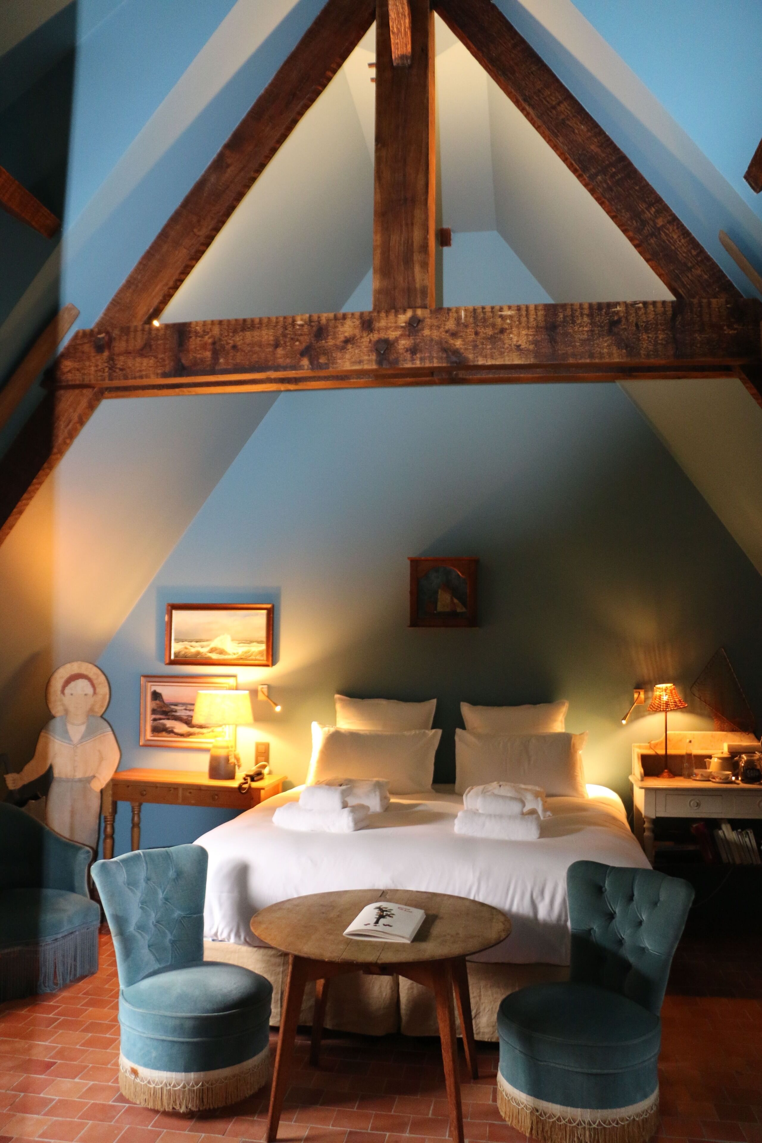 A bedroom with a sloped ceiling, a bed with white linens, two blue upholstered chairs, a small round table, and a nightstand with a lamp.