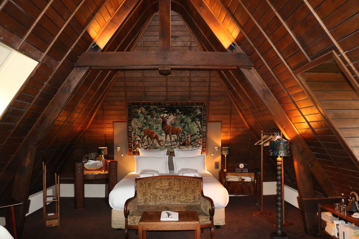 Une chambre aménagée dans les combles, avec un plafond en bois en forme de voûte, une grande peinture représentant des cerfs, un lit recouvert de draps blancs, une chaise en bois et un bureau avec une lampe.