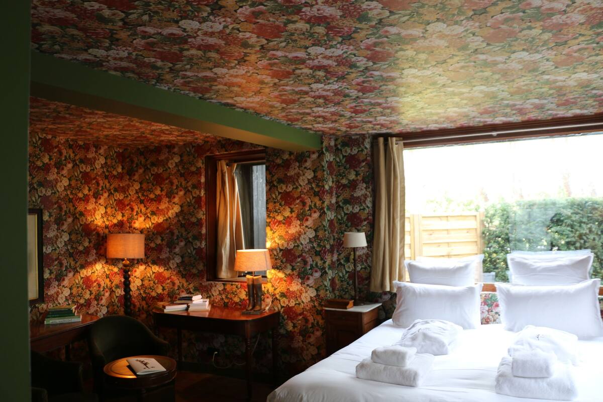 Une chambre d’hôtel avec un papier peint à motifs floraux, un lit avec du linge de maison blanc, deux lampes de chevet et une fenêtre avec des rideaux.