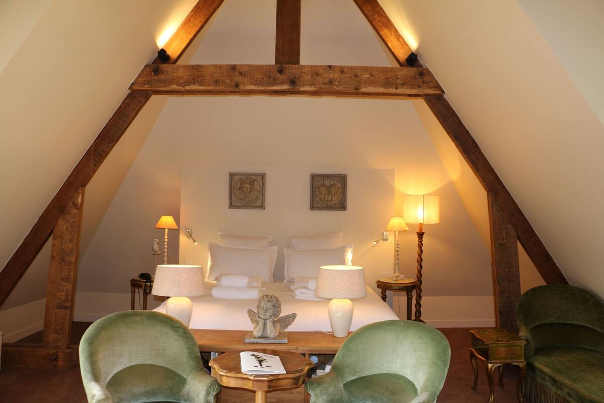 Une chambre avec un plafond en pente, des poutres en bois, un lit recouvert de draps blancs, deux lampes de chevet et une petite table sur laquelle se trouvent un livre et une figurine.