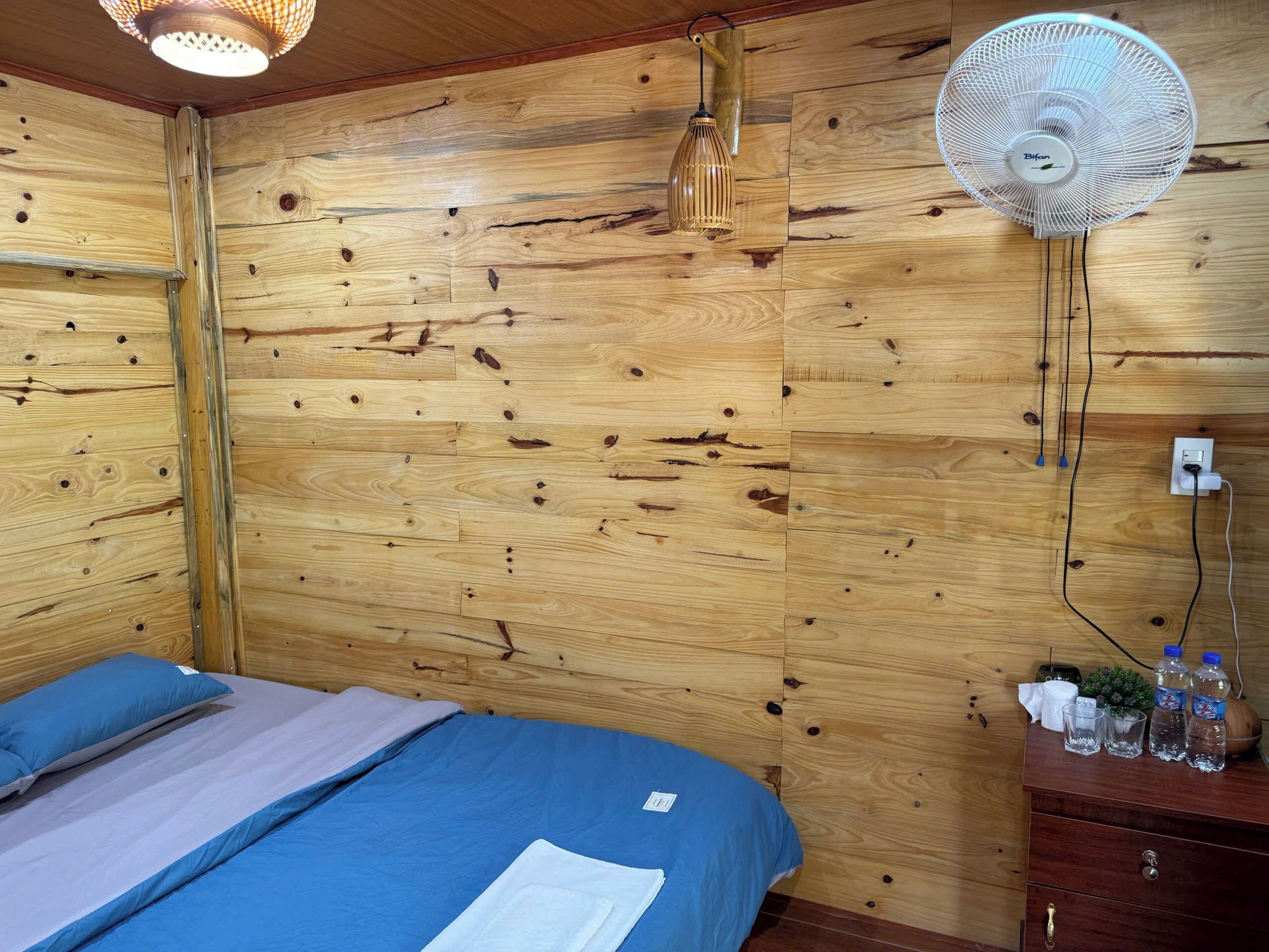 Um quarto com painéis de madeira, uma cama, um ventilador na parede e uma cômoda com garrafas e copos.