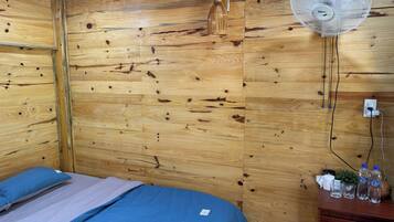 Um quarto com painéis de madeira, uma cama, um ventilador na parede e uma cômoda com garrafas e copos.