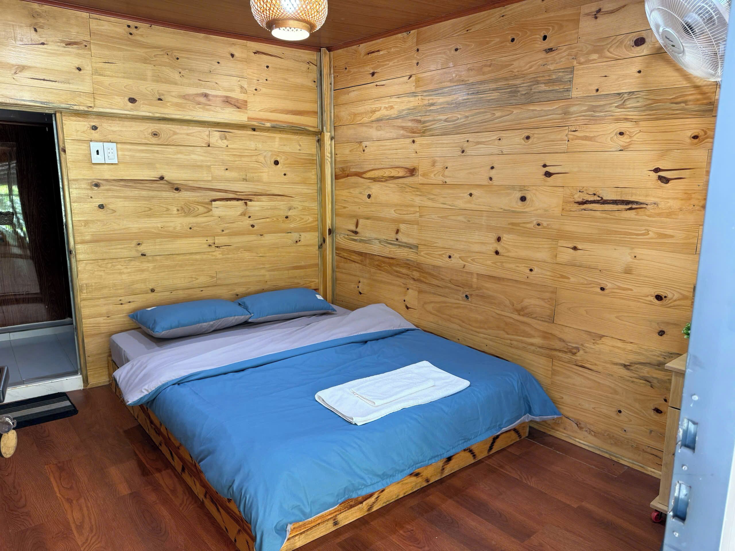 A wooden-paneled room with a bed, a fan, and a door.