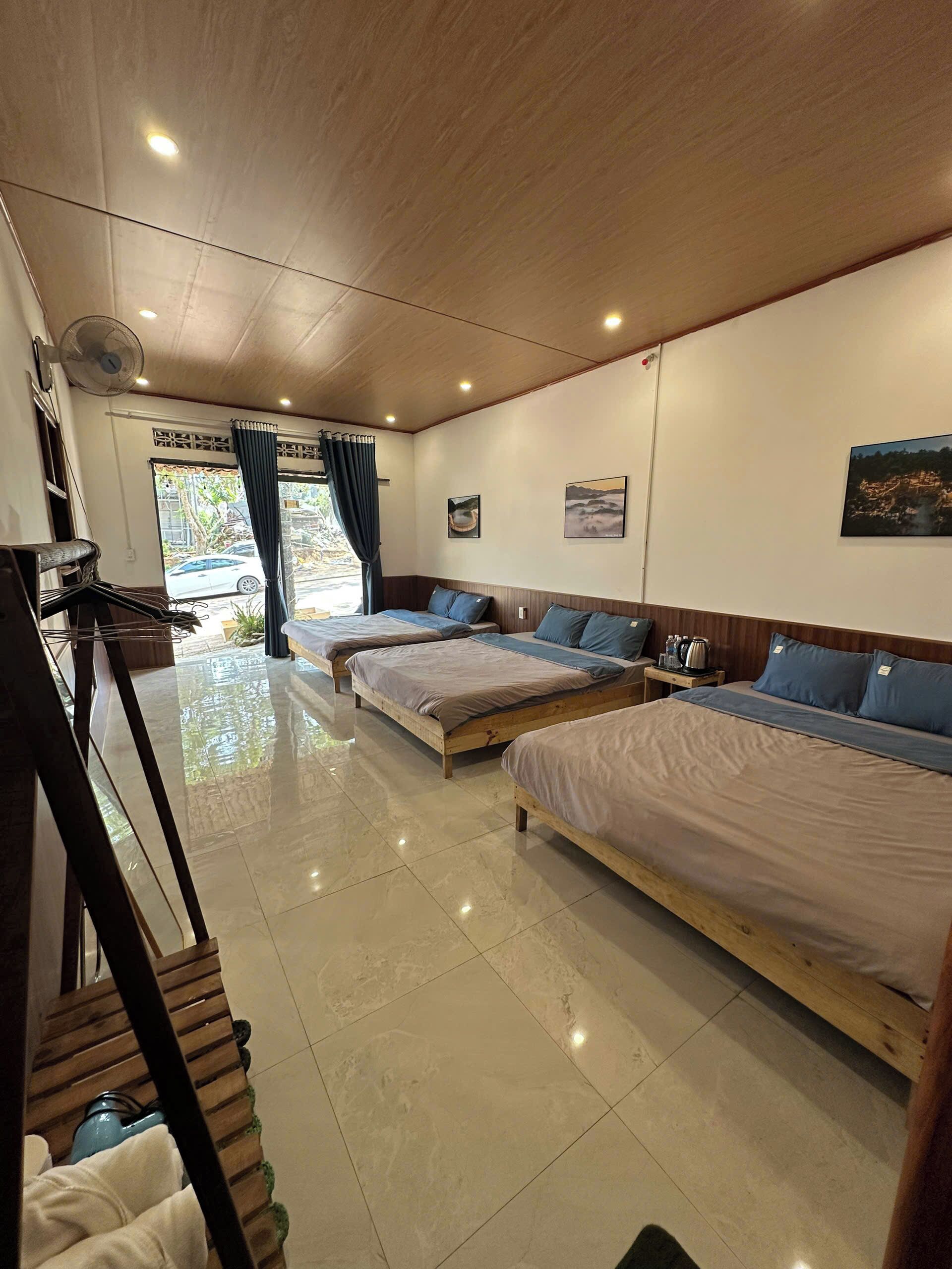 A hotel room with three beds, a fan, a window with curtains, and a picture on the wall.