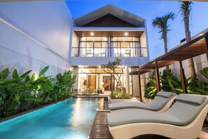 Una casa moderna de dos pisos con piscina, sillas de descanso y plantas tropicales.
