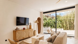 A modern living room with a sofa, a television, a cabinet, and a view of a garden. - Boho Retreat 3BR Enclosed Lounge 5min Beach (Canggu)