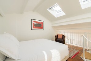 A bedroom with a bed, a framed picture on the wall, a chair, and a view of the balcony.