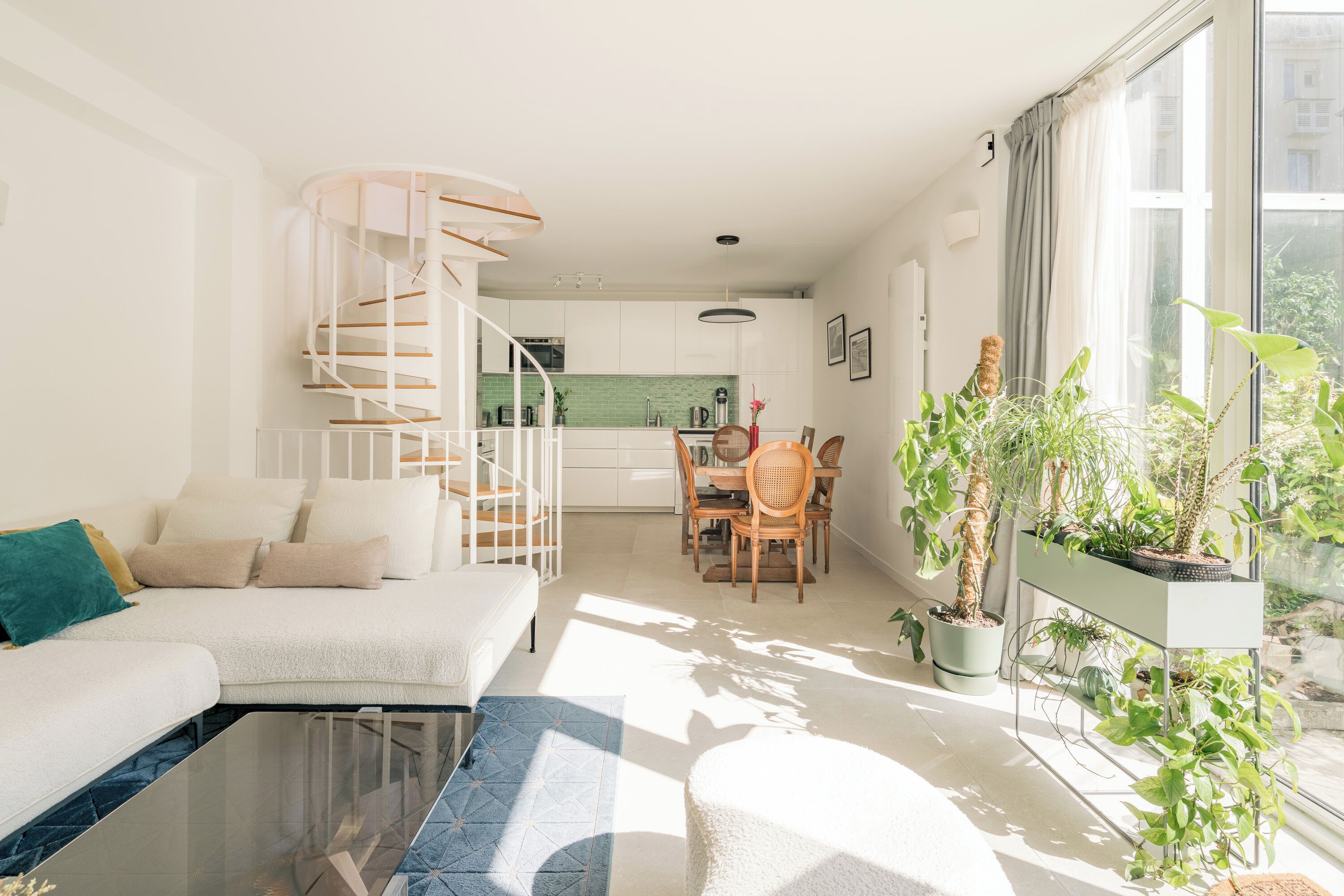 Un salon moderne avec un canapé blanc, une table basse en verre et un escalier en colimaçon menant à un niveau supérieur. On y trouve également un coin repas avec une table et des chaises en bois, ainsi qu’une grande fenêtre ornée de plantes en pot sur le rebord.