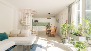 A modern living room with a white sofa, a glass coffee table, and a spiral staircase leading to an upper level. There is a dining area with a wooden table and chairs, and a large window with potted plants on the windowsill.