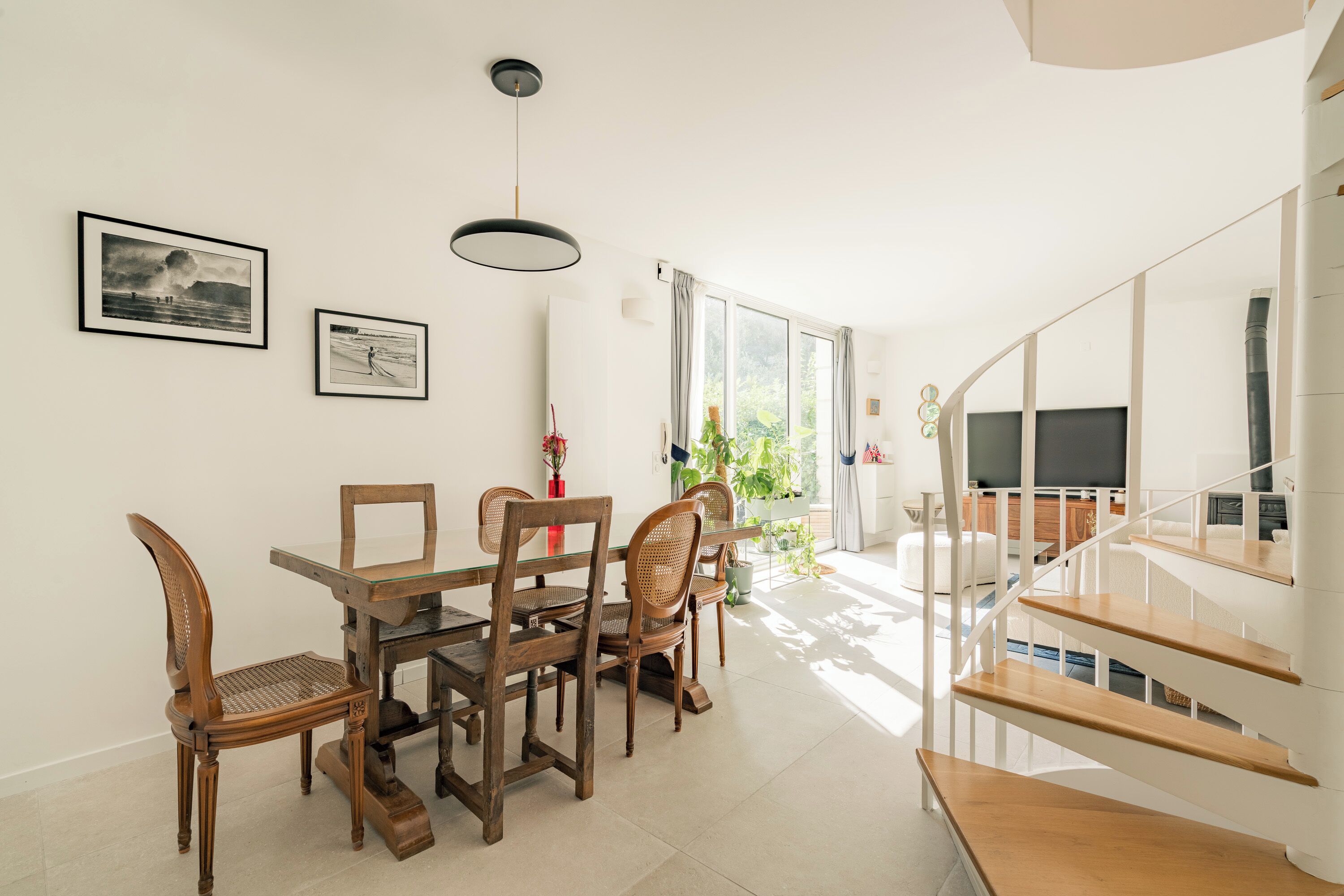 A dining area with a glass table and wooden chairs, a staircase with wooden steps, and a TV in the background.