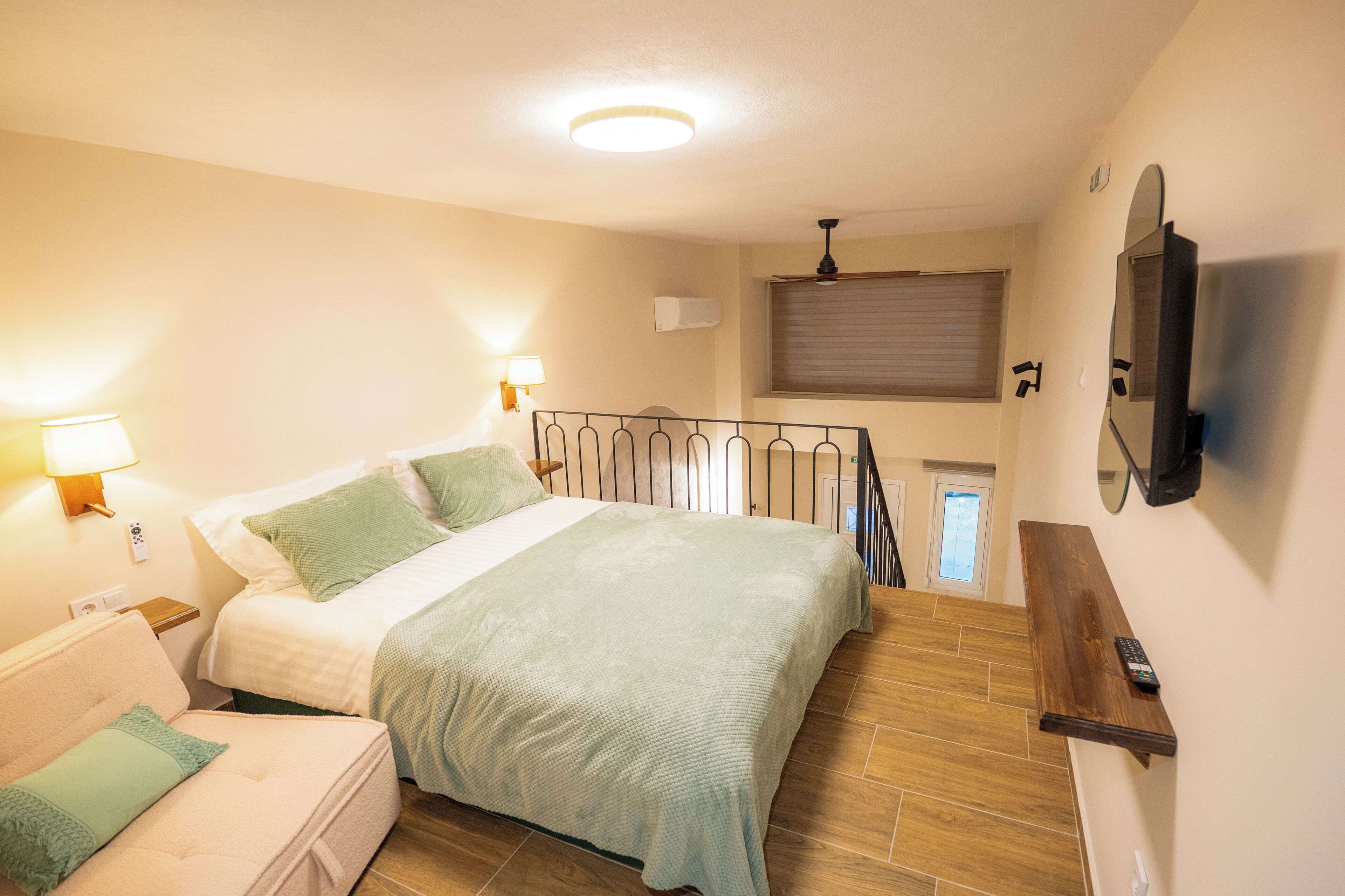 A bedroom with a bed, bedside tables, a nightstand, a sofa, a TV mounted on the wall, and a window with blinds.