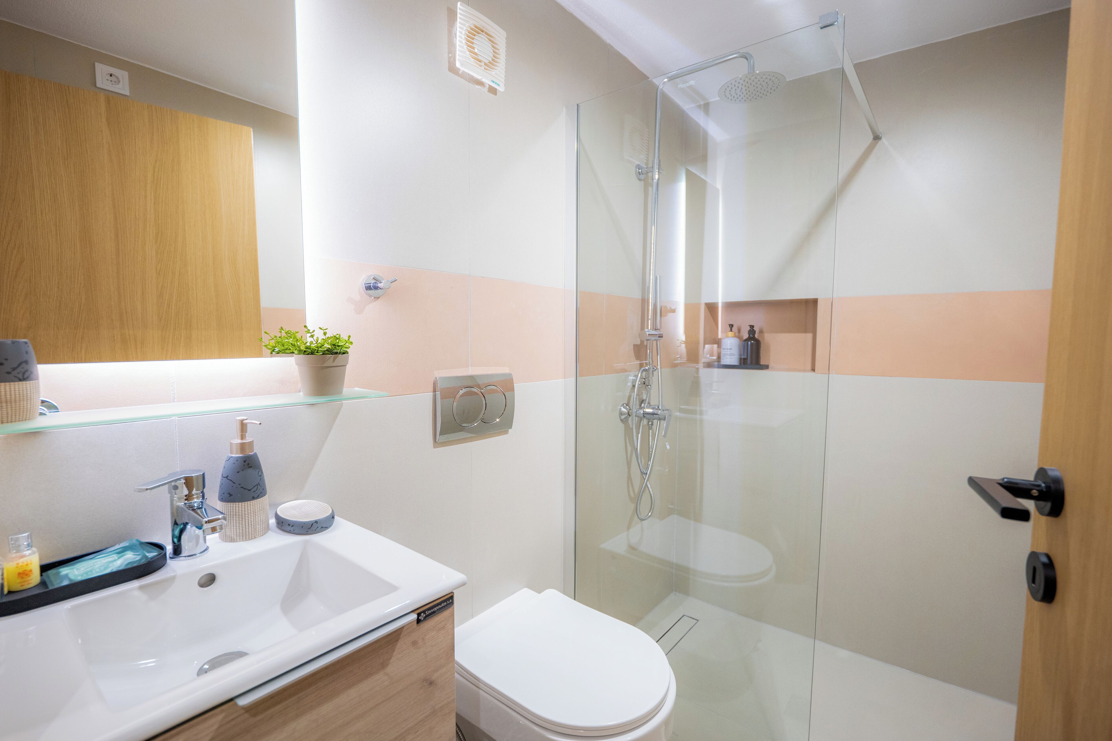 Un baño moderno con ducha envuelta en vidrio, un lavamanos blanco con dispensador de jabón y un mueble de tocador de madera.