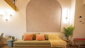 A cozy living room with a green sofa, orange cushions, a yellow throw blanket, and a large wall-mounted mirror. - Vayas Residences Agora (Trikala)