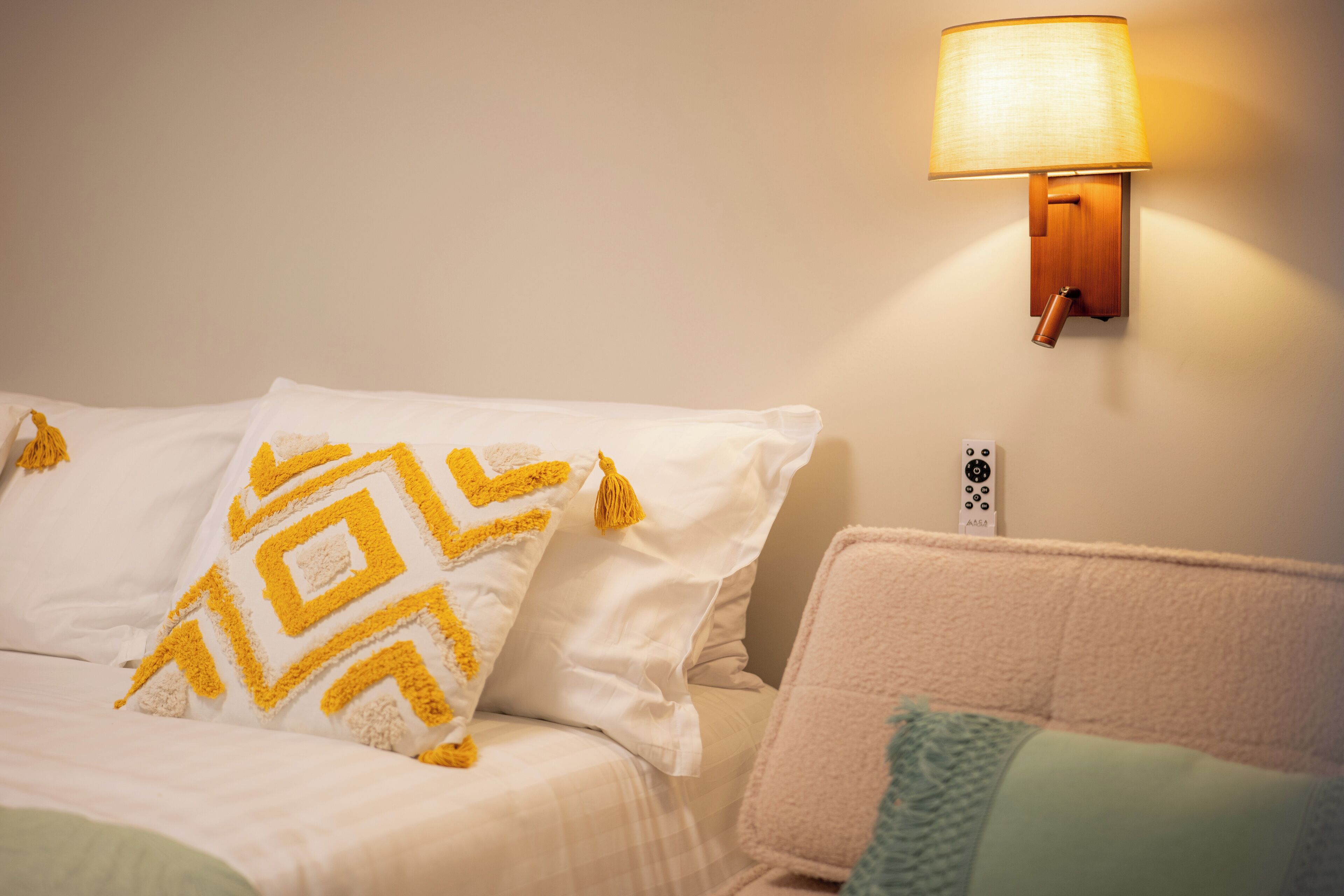 Un letto rifatto con lenzuola bianche e un cuscino decorato, una lampada fissata al muro e una presa di corrente sulla parete.