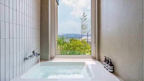 Une salle de bain moderne avec une grande baignoire, une fenêtre donnant sur un paysage verdoyant et des bâtiments, ainsi que des articles de toilette disposés sur le côté.