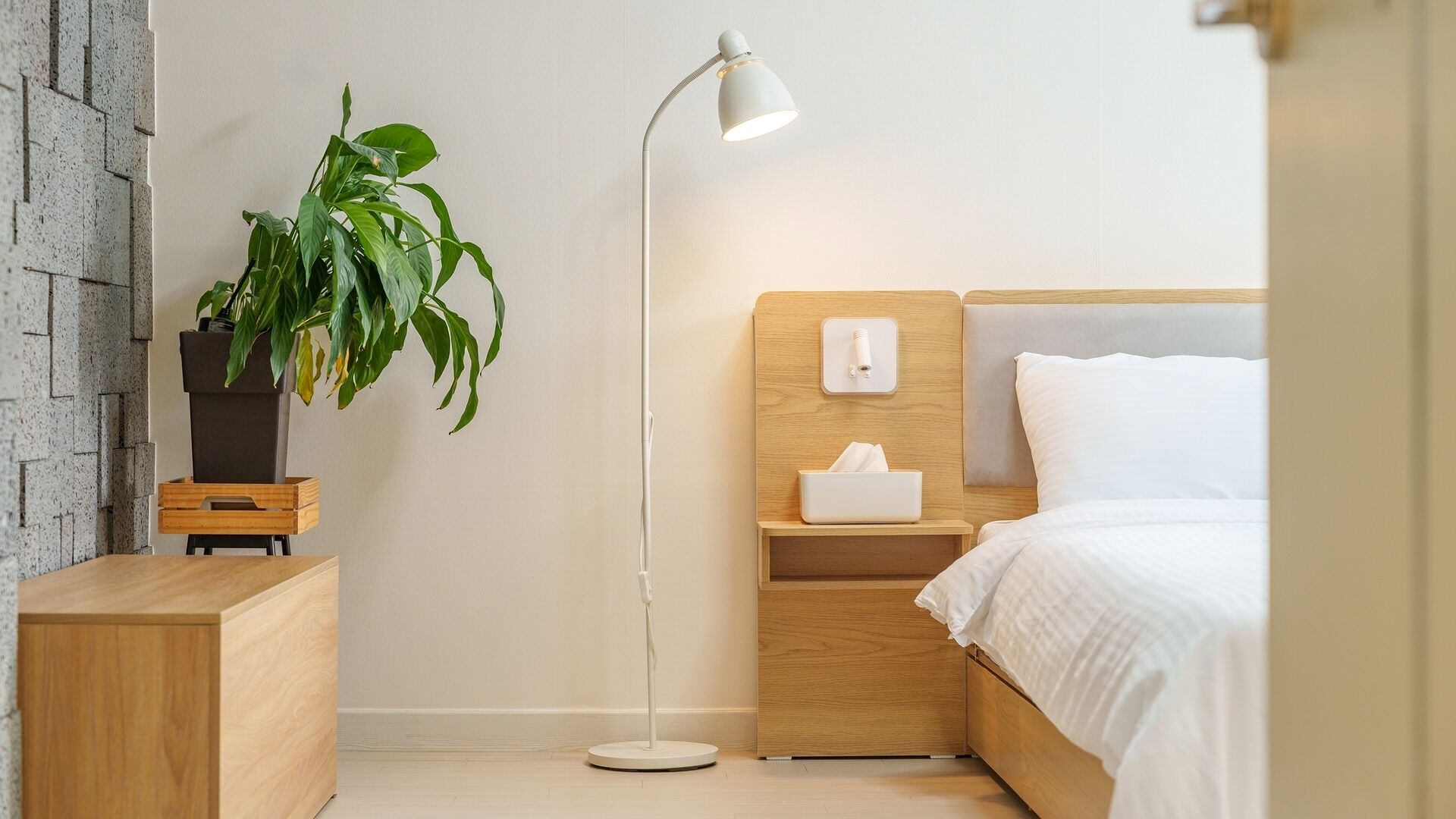 A modern bedroom with a wooden headboard, a bedside table with a lamp, a nightstand with a tissue box, and a potted plant.