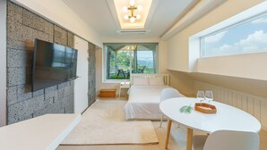 Une chambre d’hôtel moderne dotée d’une grande télévision, d’un coin repas avec une table et des chaises, d’une fenêtre donnant sur un espace verdoyant et d’un luminaire au plafond.
