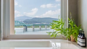 Une vue sur un pont enjambant une rivière, avec des montagnes en arrière-plan, telle qu'elle apparaît à travers une fenêtre ornée d'une plante et de bouteilles sur le rebord.