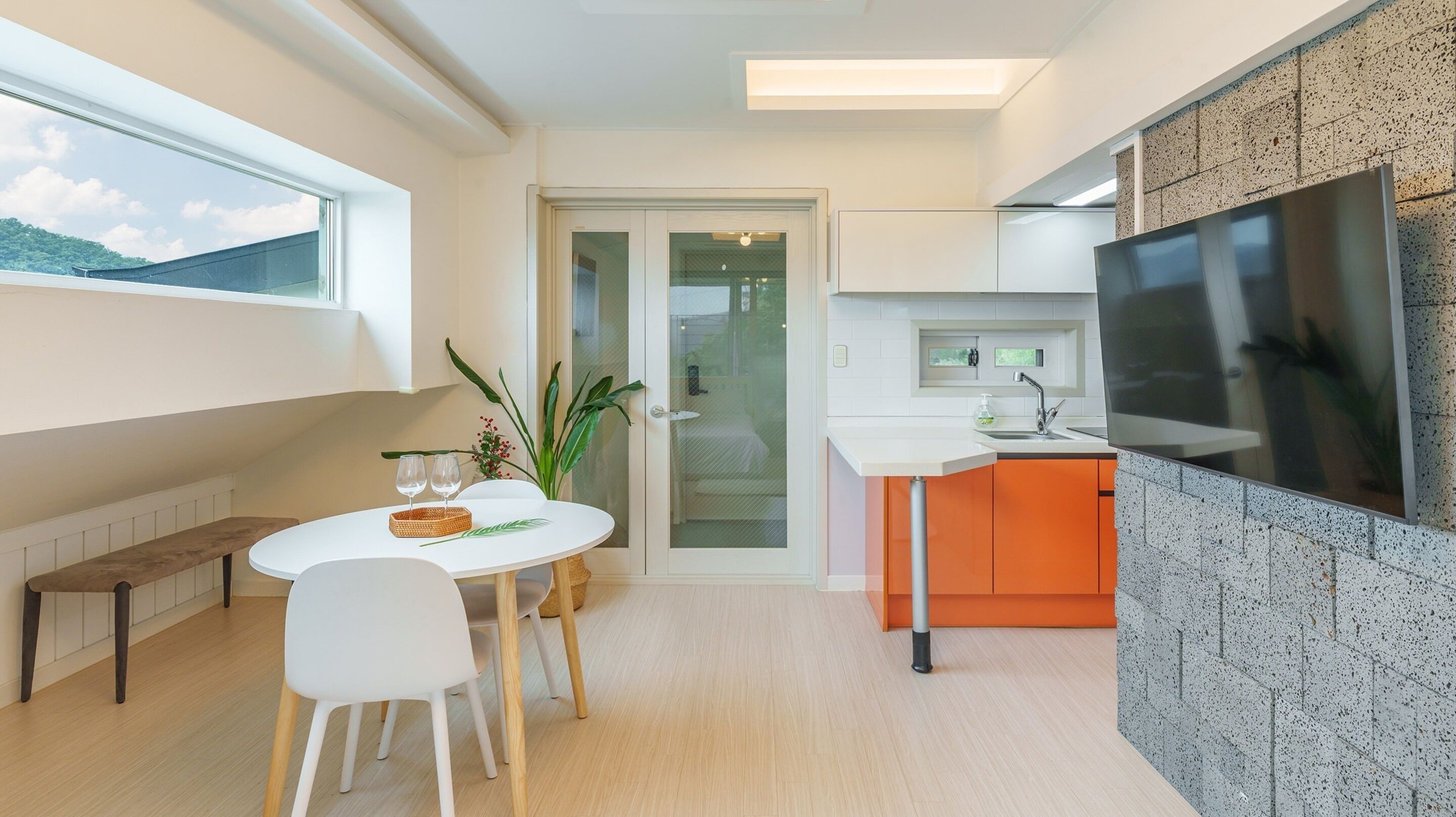 A modern kitchen with a large TV, a dining area with a bench, and a view of the outdoors.