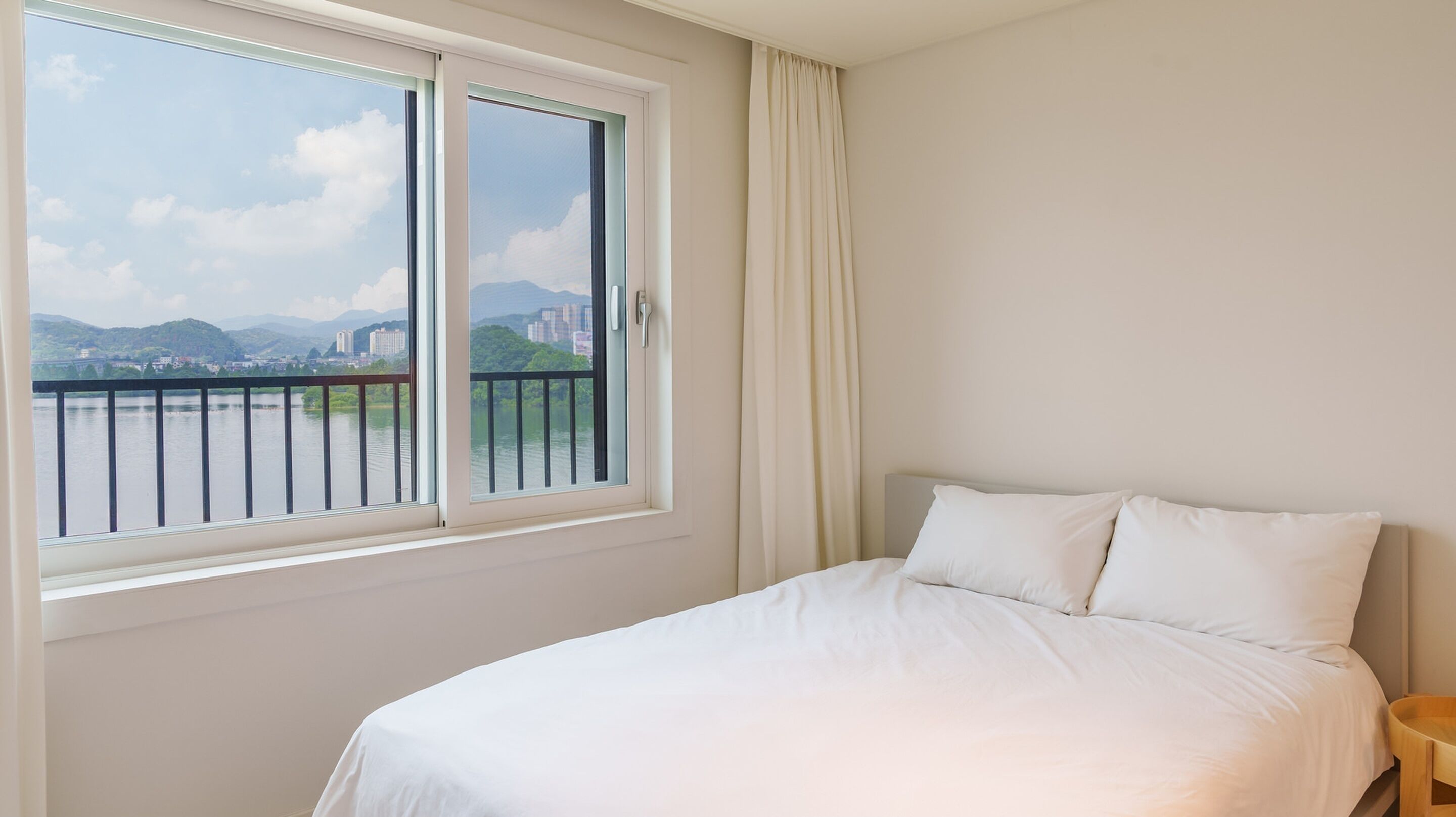Habitación de hotel con cama, ventana que ofrece vistas a un paisaje urbano y montañas, y balcón con barandilla.