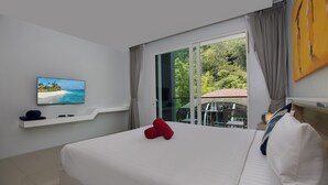 A hotel room with a bed, a television on the wall, a window with curtains, and a view of greenery outside.