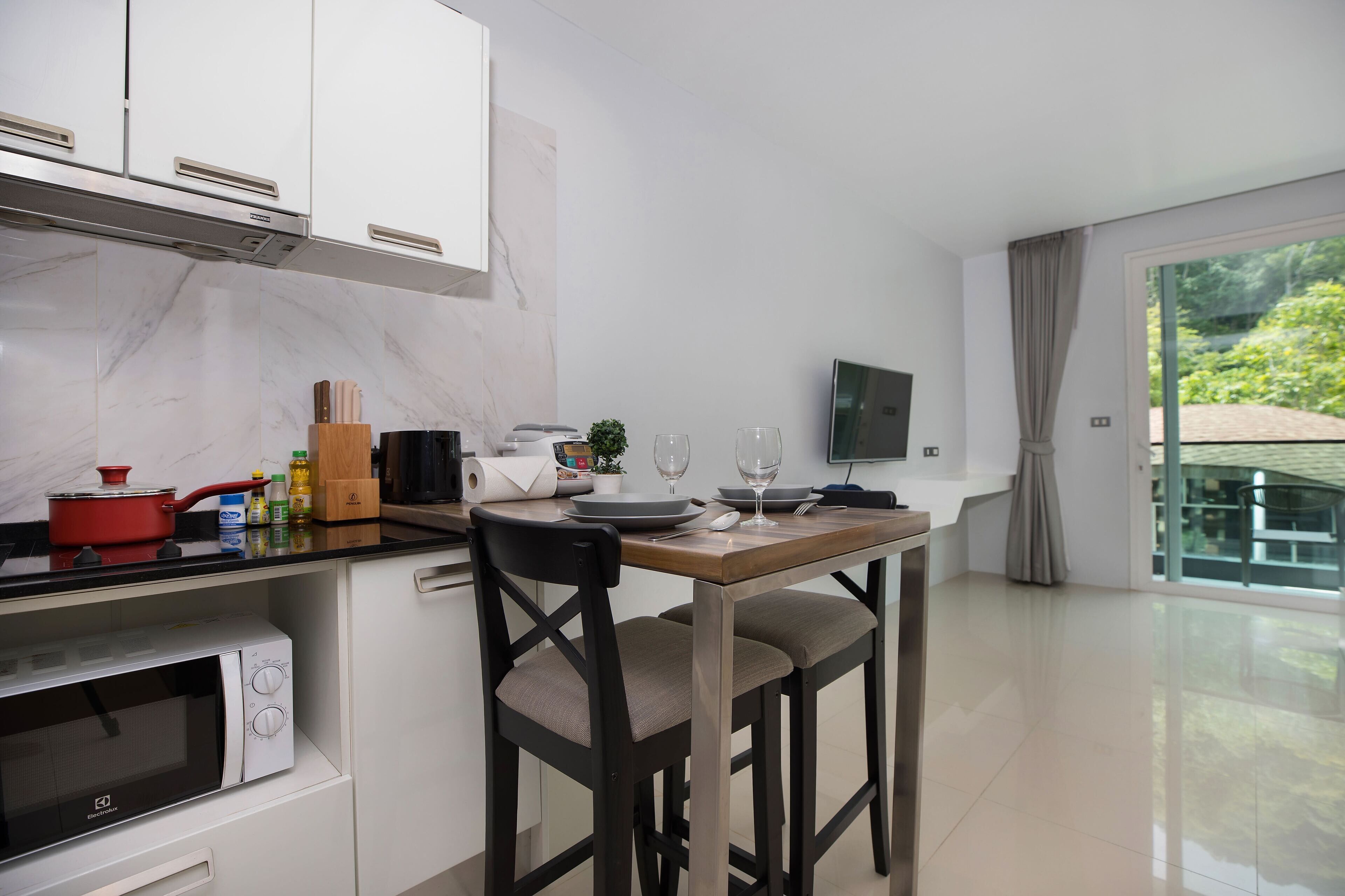 A modern kitchen with white cabinets, a built-in microwave, a wooden dining table with two chairs, and a large window offering a view of greenery.