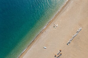 En vy över en strand sedd från luften, med klart vatten, en sandstrand och flera solstolar uppställda i en rad.