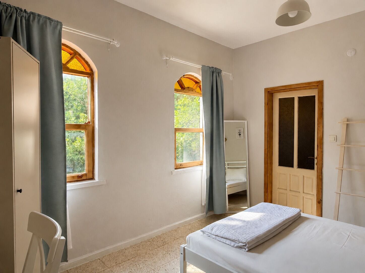 Une chambre à coucher avec un lit, deux fenêtres avec des rideaux, une porte et une petite chaise blanche.
