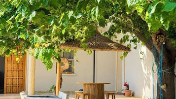 Een terras met tafels en stoelen onder een boom, een rieten dak en een houten deur.