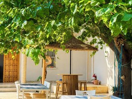 Un área con mesas y sillas al aire libre bajo un árbol, techo de paja y una puerta de madera.
