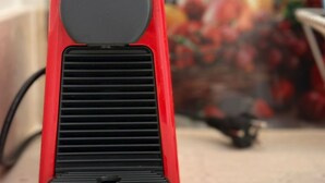 Um objeto vermelho e preto com parte superior circular e frente ventilada, possivelmente um alto-falante ou eletrodoméstico, com fundo desfocado.