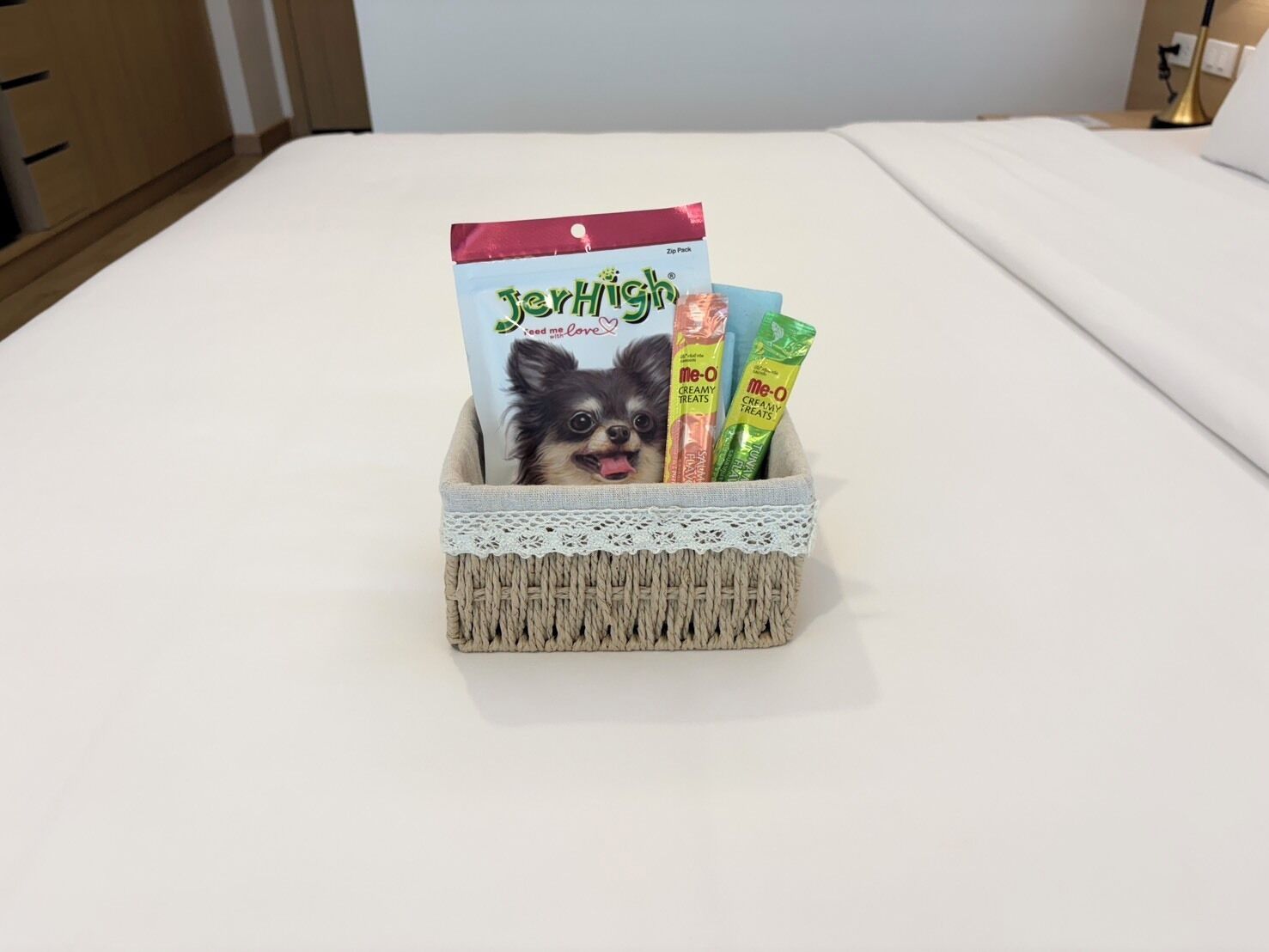 A bed with a white sheet, a wicker basket with a lace trim containing a dog food bag, and two dog treat packages.