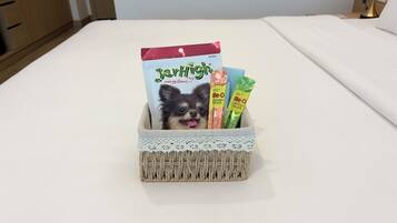 A bed with a white sheet, a wicker basket with a lace trim containing a dog food bag, and two dog treat packages.