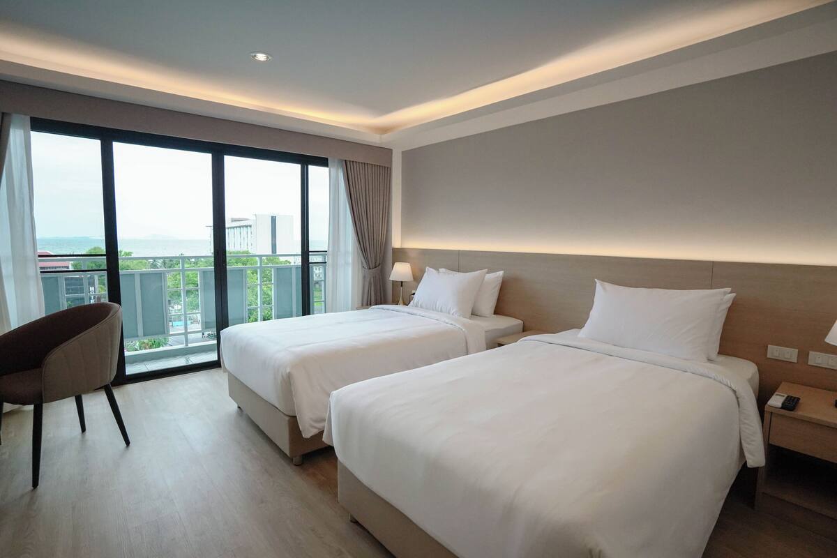 Ein Hotelzimmer mit zwei Betten, einem Sessel, einem Nachttisch und Blick auf die Stadt durch große Fenster.