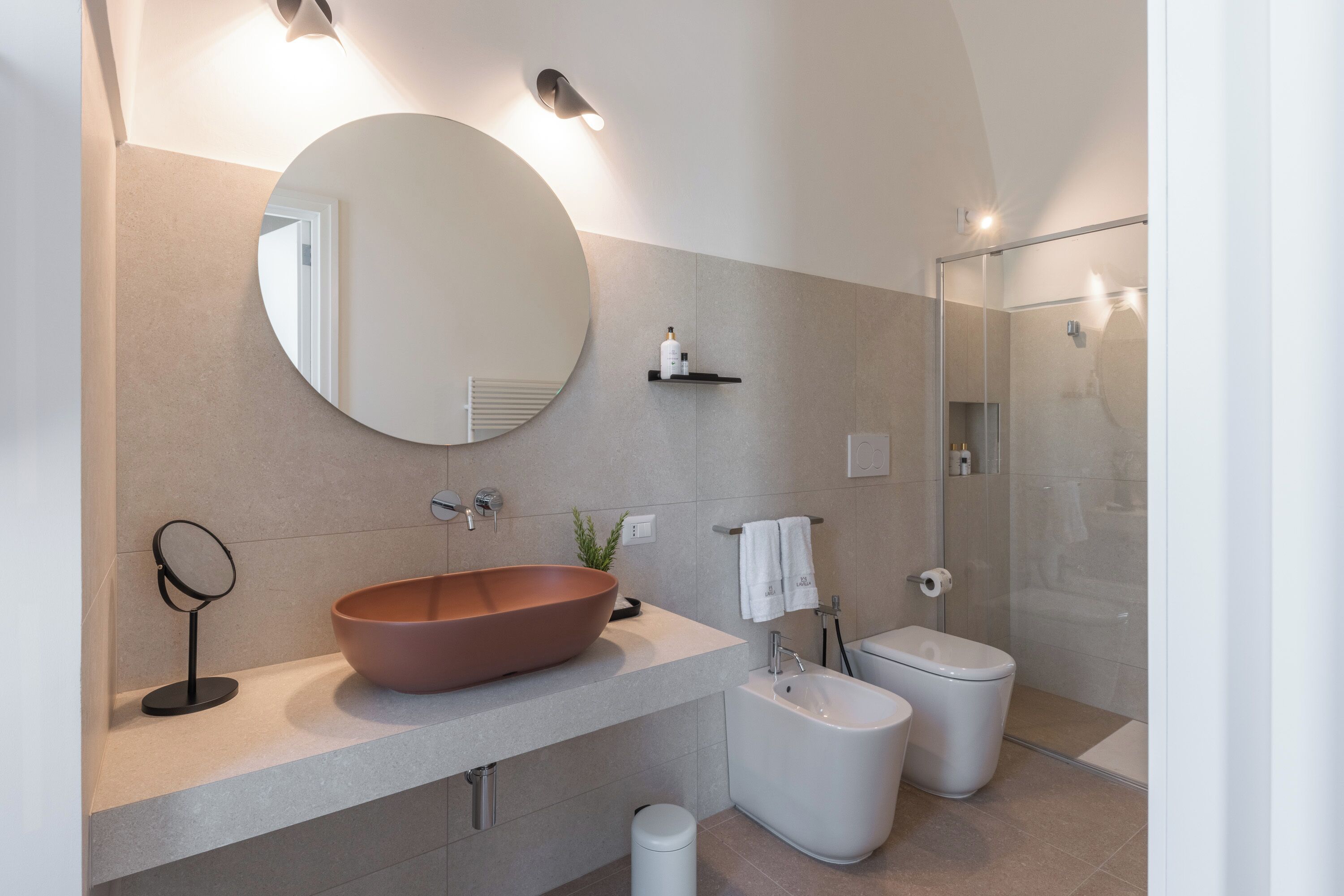 Une salle de bain moderne avec un grand miroir rond, un vasque imposant, une baignoire sur pieds et une douche à parois de verre.