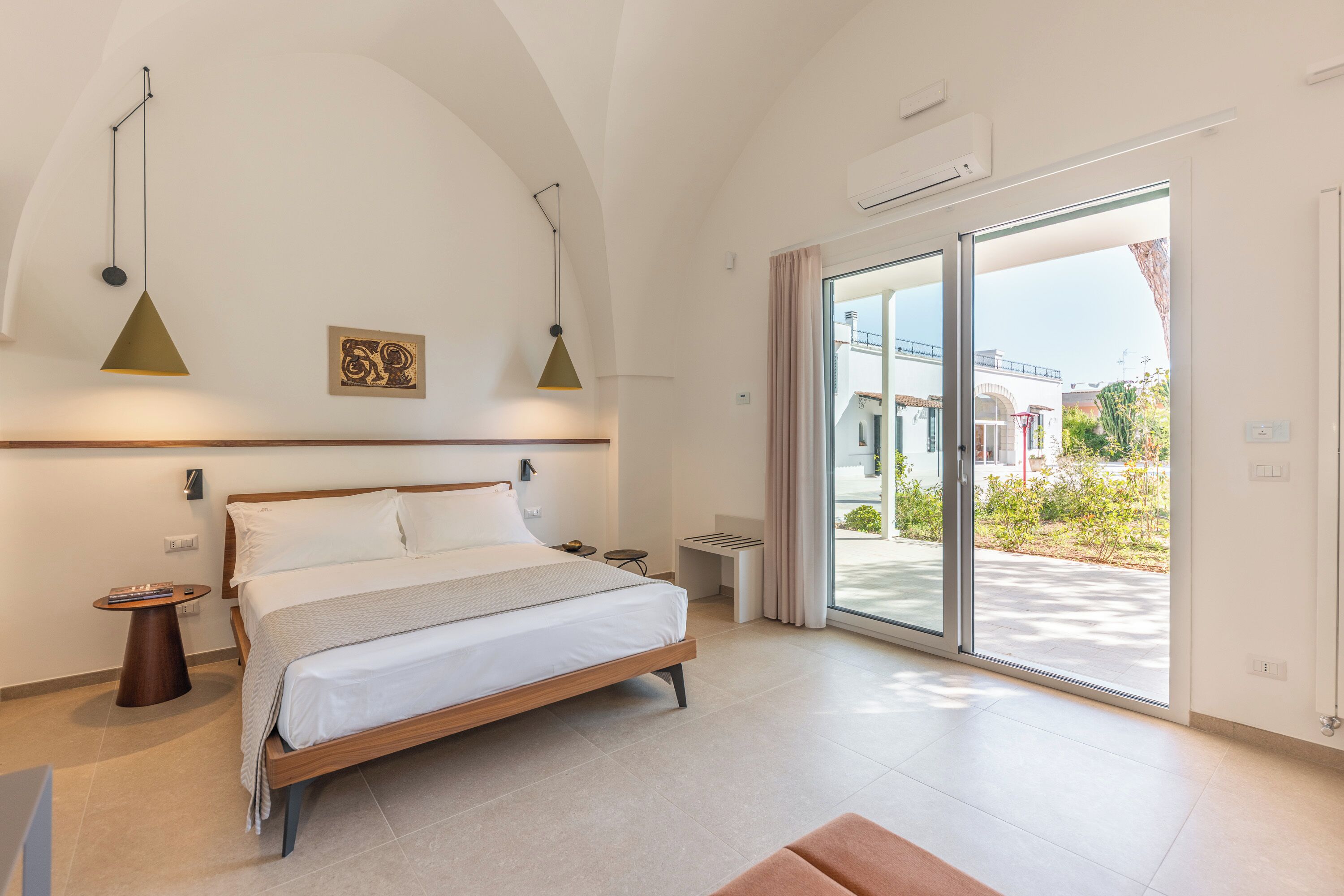 Een moderne slaapkamer met een bed, nachtkastjes, een hanglamp en uitzicht door een schuifdeur naar buiten.