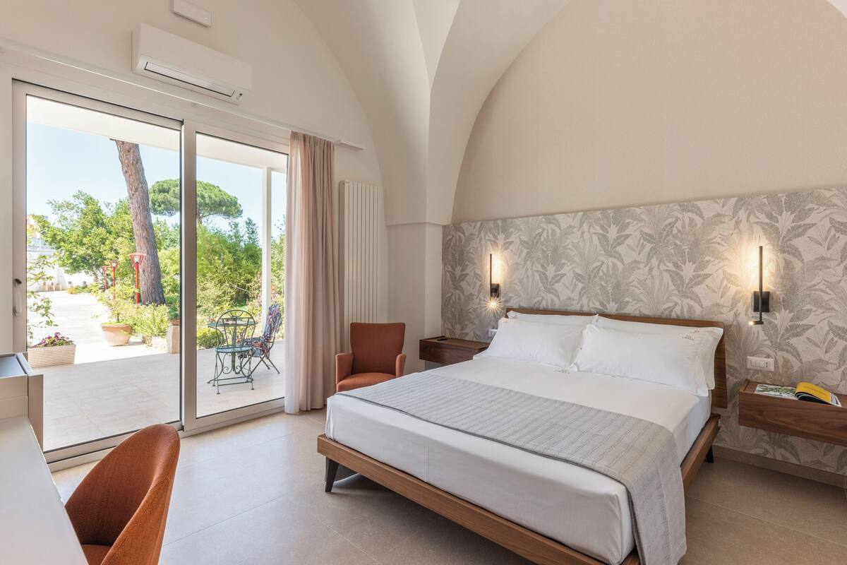 A bedroom with a large bed, a chair, a nightstand, and a view of the outdoors.