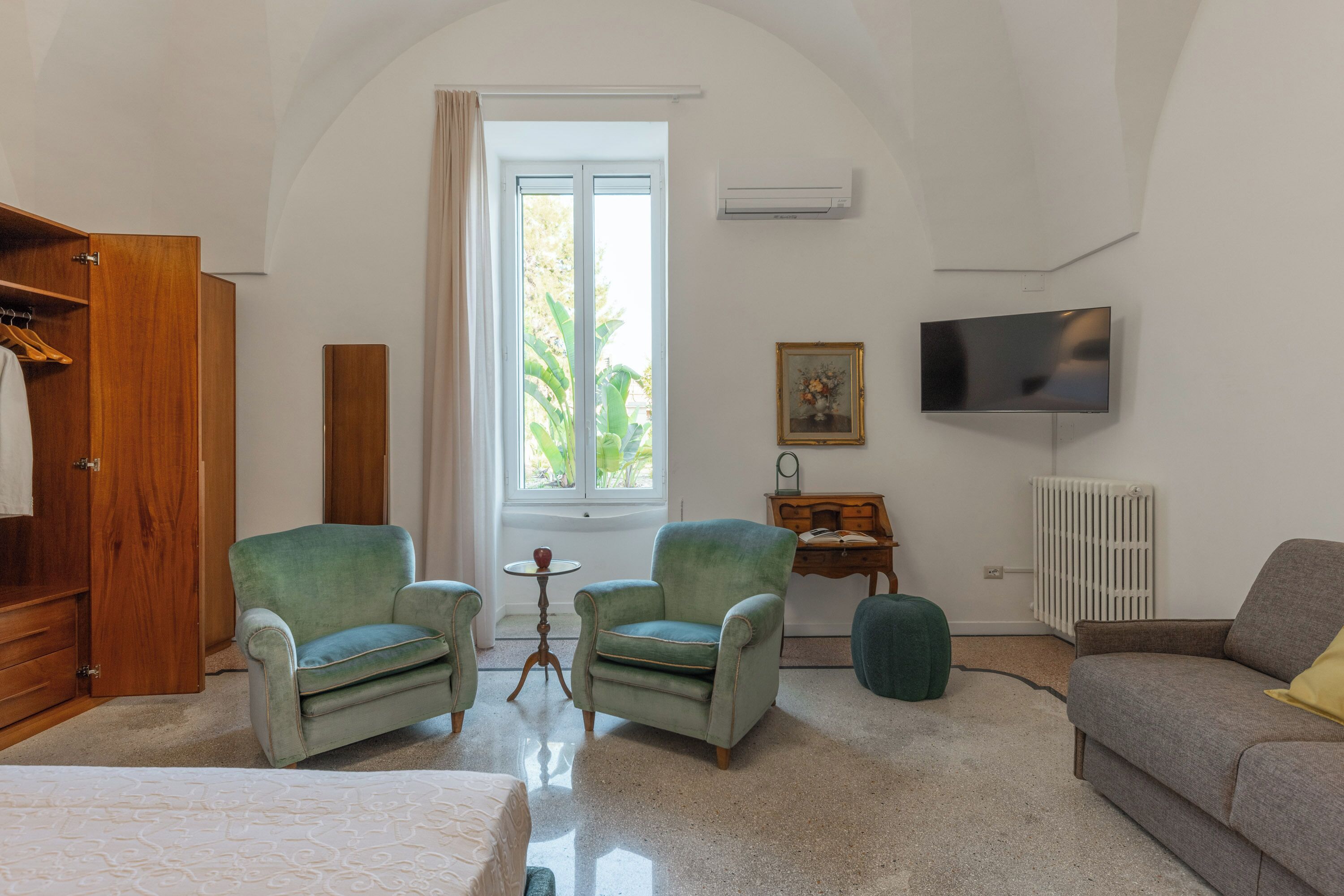 Une pièce comprenant un lit, deux fauteuils, un canapé, un téléviseur, un radiateur et une fenêtre avec des rideaux.
