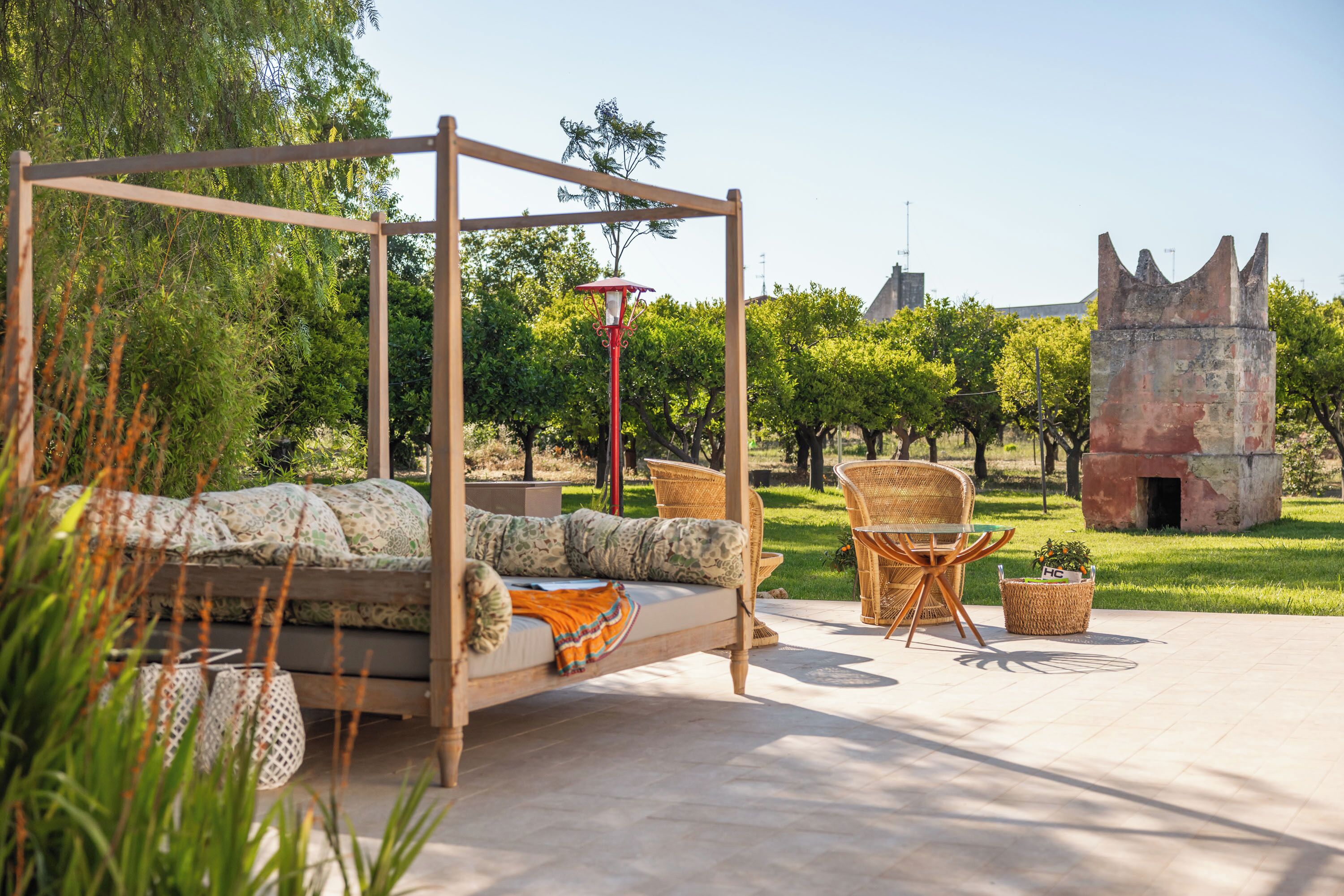 A wooden four-poster bed with a red lamp, wicker chairs, and a stone structure in the background.