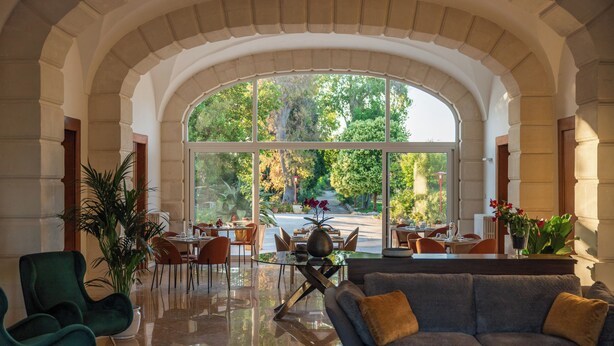 Un vestíbulo amplio con un gran ventanal de arco que ofrece vistas al exterior, un área de descanso con sofá y sillones, y un comedor con mesas y sillas.