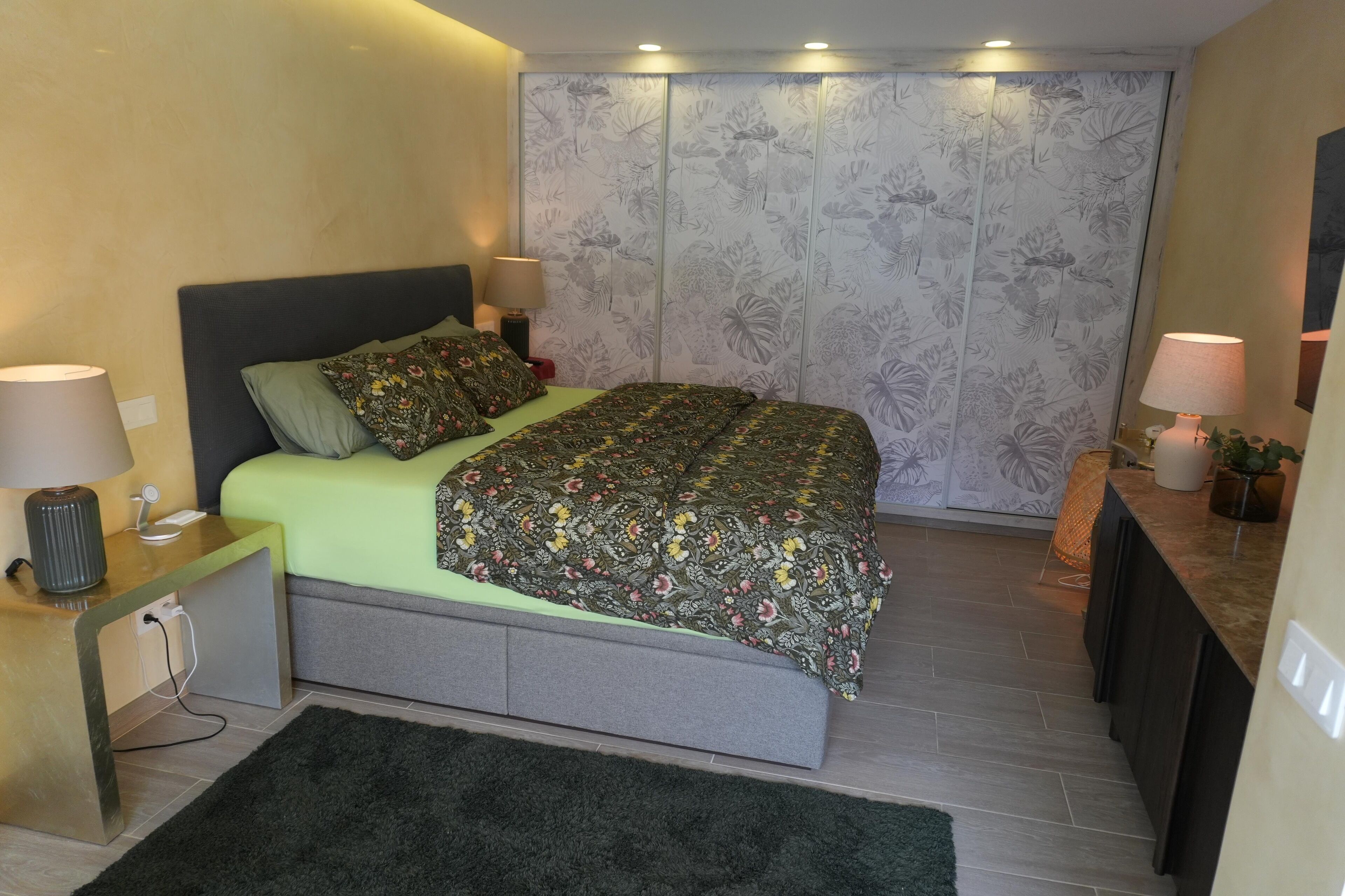 Un dormitorio con cama, mesitas de noche, cómoda y un panel decorativo con estampado floral.